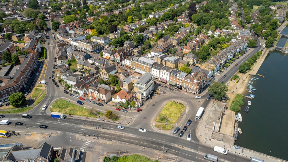 An aerial view of Richmond upon Thames showing a mix of residential buildings, including terraced houses and apartment complexes, situated along streets lined with trees. In the foreground, a busy road with cars, buses, and parked vehicles runs parallel to a river with a paved promenade. Nearby, a small roundabout surrounds a grassy patch with benches, while the riverbank features moored boats and a pedestrian walkway. The scene is illuminated by natural daylight, emphasizing the clean, well-maintained surfaces and lush greenery, reflecting a typical urban landscape that benefits from professional surface cleaning and maintenance services through companies like [COMPANY_NAME].