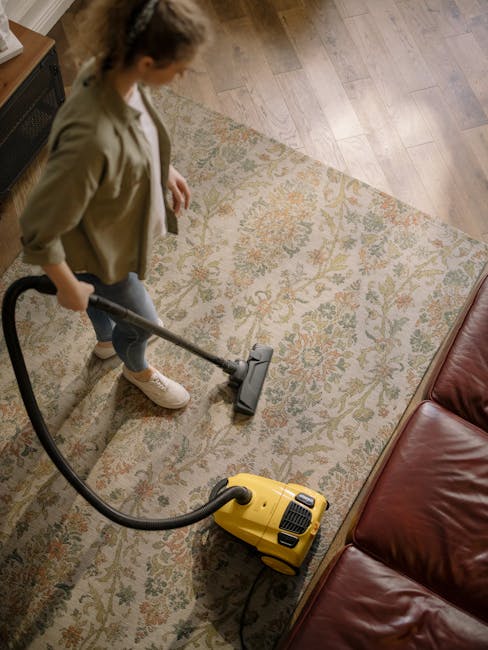 A woman performing surface cleaning with a vacuum cleaner on a patterned carpet in a living room. The room features a wooden floor border and a red leather sofa, with natural light illuminating the space. She is dressed casually in a beige jacket, grey jeans, and white sneakers. The carpet, which appears clean and well-maintained, has a floral design with beige, green, and orange hues. The vacuum cleaner has a yellow body and a black hose, and she is holding the handle as she vacuums the carpet. This scene exemplifies deep cleaning and maintenance practices, consistent with local cleaning services, including those provided by [COMPANY_NAME], to ensure hygienic and pristine surfaces in residential settings.