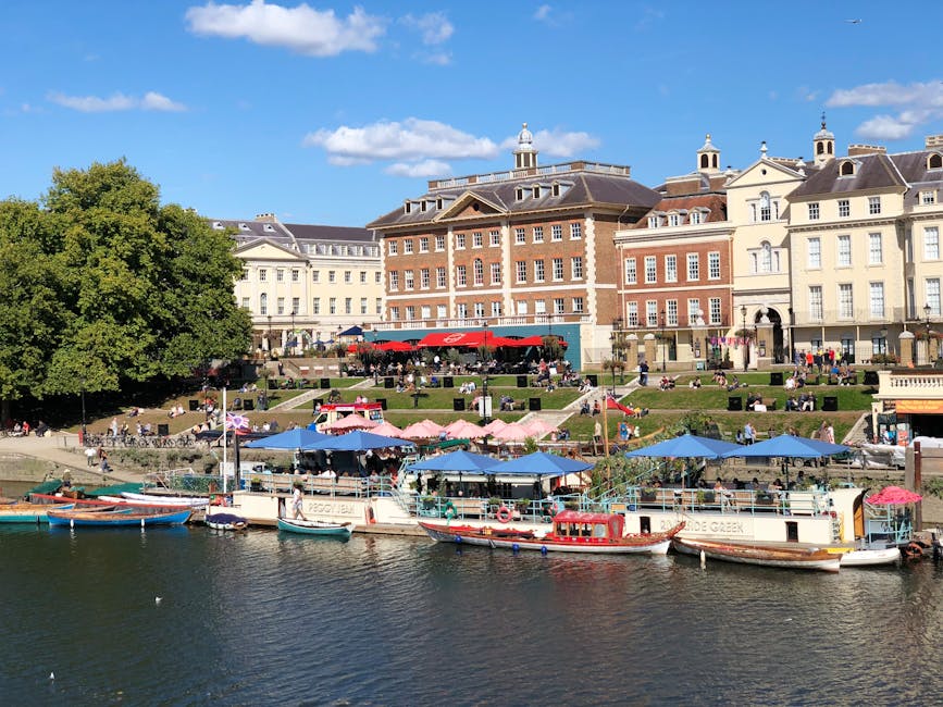 An aerial view of Richmond upon Thames showing a mix of residential buildings, including terraced houses and apartment complexes, situated along streets lined with trees. In the foreground, a busy road with cars, buses, and parked vehicles runs parallel to a river with a paved promenade. Nearby, a small roundabout surrounds a grassy patch with benches, while the riverbank features moored boats and a pedestrian walkway. The scene is illuminated by natural daylight, emphasizing the clean, well-maintained surfaces and lush greenery, reflecting a typical urban landscape that benefits from professional surface cleaning and maintenance services through companies like [COMPANY_NAME].